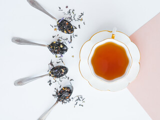 A cup with tea and a saucer stands on the corner of a beige sheet. Dry black tea mixed with herbs for brewing is poured into spoons, diverges from the cup in rays. Layout for tea business.