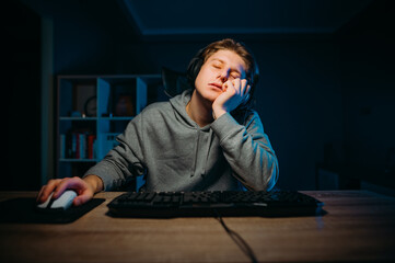 A sleepy gamer sleeps at the computer and falls asleep while bored playing in a dark room at home.