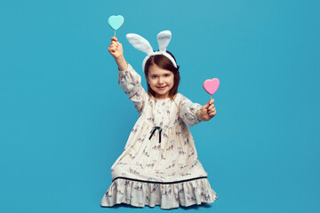 Cute kid girl, wearing easter bunny ears, having two cookies in heart shape on sticks, isolated over blue background