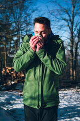 Attractive young man enjoying cup of hot tea outdoors in snowy forest  on cold but sunny winter morning. Holding hot beverage with a joy  on his face and eyes closed, trying to warm up.