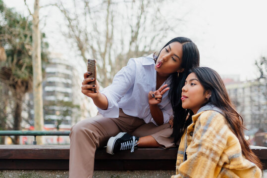 Two Latin girls sitting in the street taking pictures of themselves making faces with a phone. Sisters making a funny selfie for their family.