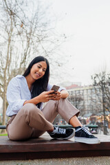 young latin girl sitting in a public city park using an online messaging app from her smart phone.