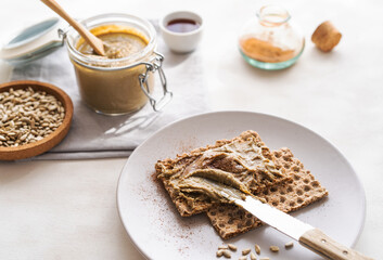 Homemade sunflower seed spread in jar with roasted seeds, maple syrup, cinnamon, crisp bread on light background