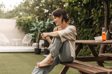Naklejka premium Portrait of beautiful young caucasian woman smiling while sitting on bench with mobile phone. Dark-haired model in casual light clothing. Social media addiction concept