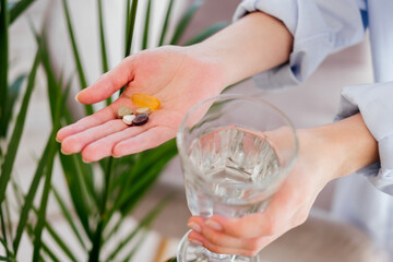 Female hands holding pills, vitamins capsules and glass of water in front of leaves indoors. Taking medicine concept