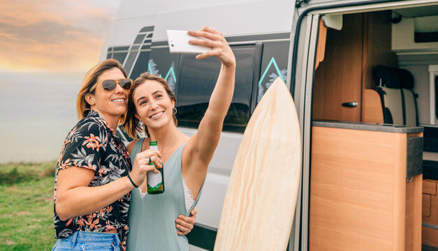 Two happy young women taking a selfie with their cell phone next to their camper van during a trip