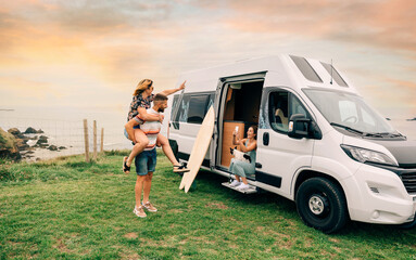 Young woman taking a photo with her cell phone of her friends having fun piggybacking next to their camper van during a trip - obrazy, fototapety, plakaty