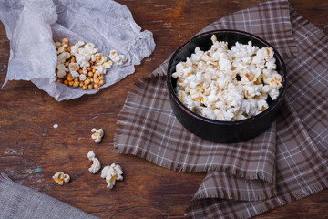 Heap of popcorn in black bowl on wooden rustic background. Some popcorns on fabric, paper and table