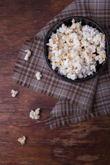 Heap of popcorn in black bowl on wooden rustic background. Some popcorns on fabric and table....