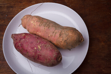 Orange, purple raw sweet potatoes, yam on white plate, wooden rustic table. Farm harvest, organic vegetables, shop local vegan food