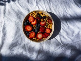On a white sheet, a cup with cereal, strawberries and blueberries. 