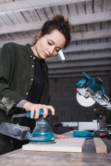 Carpenter using blurred sander on blurred plank near miter saw in workshop.