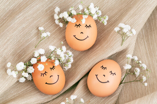 Three happy Easter eggs with cute faces in floral wreath crowns on brown burlap background.