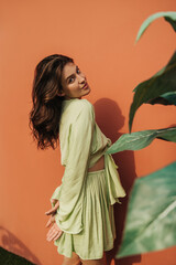 Modern young caucasian woman stands in front of red wall, holds her hands behind back, looks modestly at camera. Brunette with loose hair in light green dress. Femininity and beauty sensuality concept