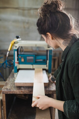 Tattooed carpenter holding blurred plank near bench thicknesser on background.