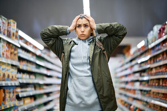 Young woman feels shocked about rising food prices while standing among the aisle with groceries in supermarket.