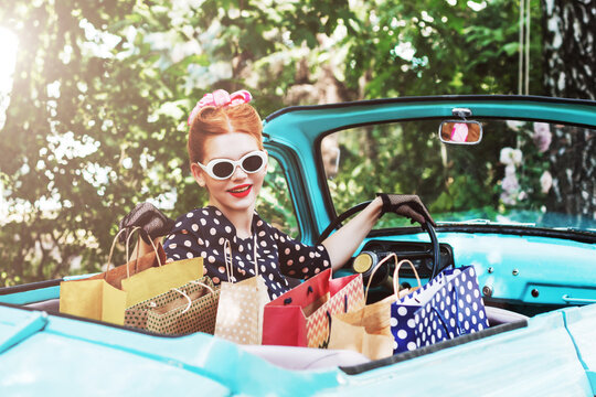 Beautiful stylish woman in vintage bright dotted clothes and glasses is driving mint old car after shopping. Fashion pin-up girl bought goods and products. Retro style concept. Black friday sale.