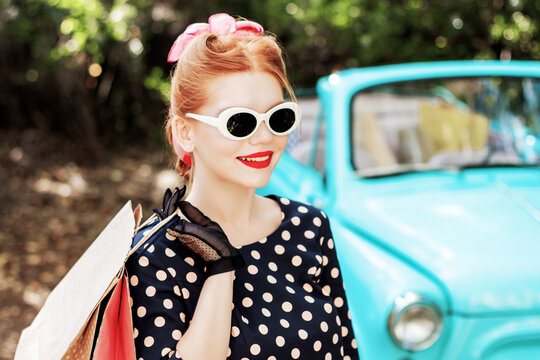 Beautiful stylish woman in vintage bright dotted clothes and glasses is holding shopping bags on background of mint old car. Fashion pin-up girl with makeup and hairstyle outdoor. Retro style concept.