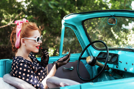 Beautiful stylish woman in vintage bright dotted clothes, sitting in mint old car. Fashion pin-up girl is doing makeup while driving. Retro style concept. Carnival, costume parties and thematic events