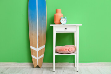 Table with vase, alarm clock and surfboard near green wall in room