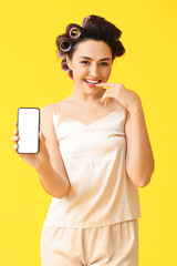 Beautiful young woman in hair curlers and with phone on yellow background