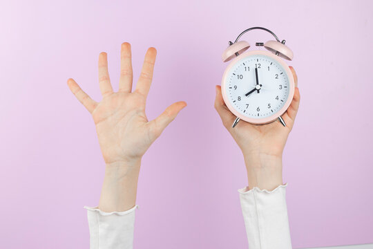 Human arm is raised up holding pink alarm clock and showing open palm, five finger on pink background.