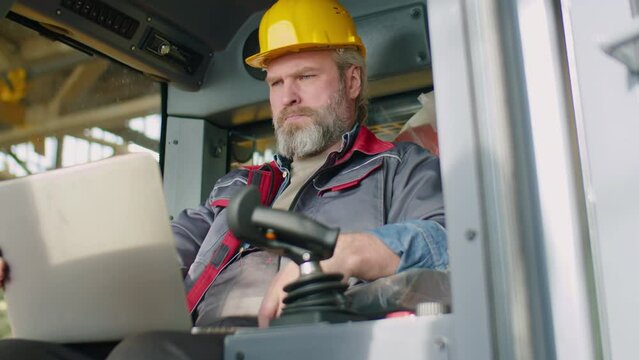 Senior maintenance technician in hardhat and workwear sitting in construction machine and using laptop while testing heavy equipment in factory
