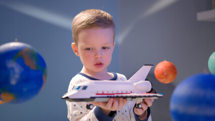 Cute boy is playing with toy space shuttle, flying in space among planets of solar system. Child...