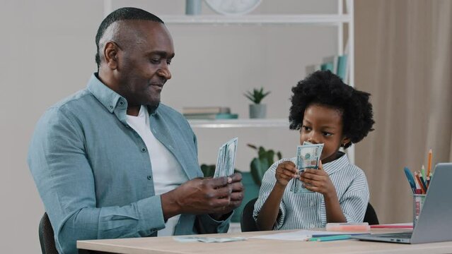 African american family sitting at desk adult father home teacher teaches financial literacy arithmetic little daughter girl learns count with help paper money dollar bills joyful child plays with dad