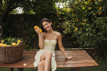 Cheerful young european lady is sitting at street table in garden, holding lemons in her hand. Girl...