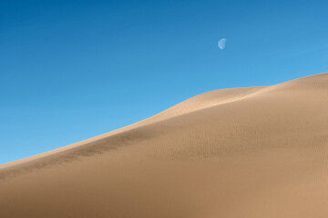 Fototapeta na wymiar Moon against blue sky over sand dunes. Sand dunes against the blue sky.