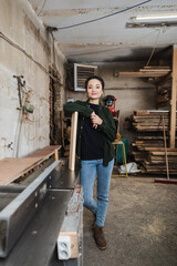 Happy woodworker looking at camera near plank and jointer machine.