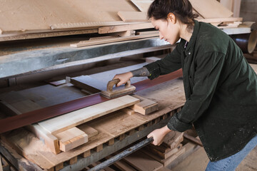 Side view of tattooed carpenter in sawdust polishing plank in workshop.