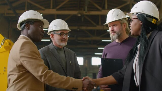 African American female engineer in hardhat and formalwear shaking hands and speaking with multiethnic male colleagues in factory