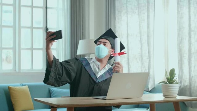 Young Asian Man Wearing Protection Face Mask, Taking A Selfie While Wearing A Graduation Gown And Cap, Hand Holding University Certificate In Living Room
