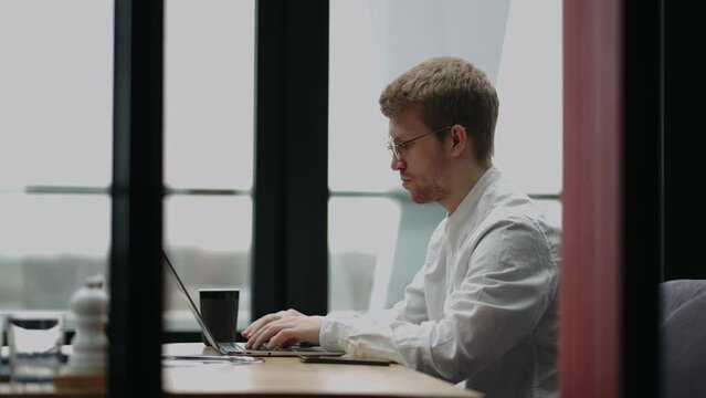 young man, wearing glasses and smiling, as he works on his laptop to get all his business done early in the morning with his cup of coffee