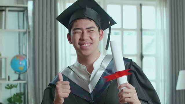 Excited Asian Man Holding A University Certificate, Smiling And Thumb Up To Camera. Male Graduate Wearing A Graduation Gown And Cap Sitting On The Living Room
