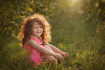 A girl in a pink dress, with beads, with long curly red hair is sitting in the grass and smiling cheerfully, the summer sun highlights her hair
