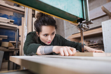 Fototapeta na wymiar Brunette carpenter holding blurred wooden board and working in band saw in workshop.