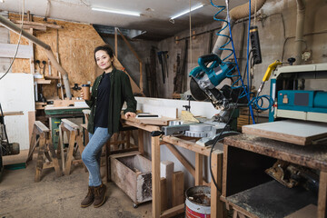 Smiling furniture designer holding coffee to go near gadgets and equipment in workshop.