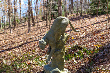 a gray statue of a panther on a hillside covered with brown fallen autumn leaves and bare winter...