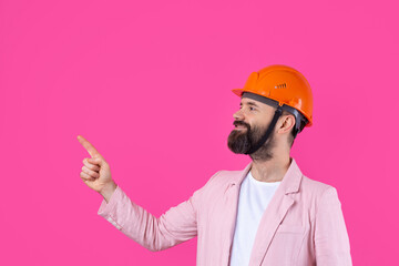 Portrait of young construction engineer wear orange hard hat, in a pink jacket standing on red studio background. The man points with his hand.