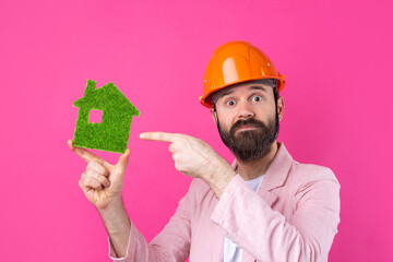 Portrait of young construction engineer wear orange hard hat, in a pink jacket standing on red studio background. A man holds a green eco house.