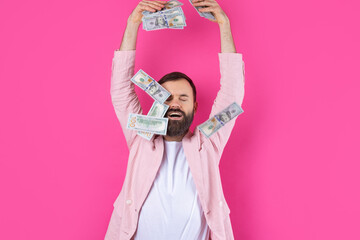 Portrait of a contented young businessman with a beard dressed in a pink jacket showing us dollar banknotes against a red studio background. Taste, smell of money.