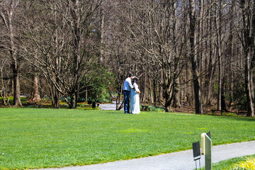 a pregnant woman wearing white and a man with a white shirt holding each other taking pictures surrounded by lush green grass, bare winter trees and lush green plants at Gibbs Gardens in Ball Ground - Powered by Adobe