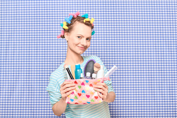 Happy young woman with colorful hair curlers is holding makeup bag