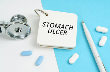 On a white and blue surface are pills, a stethoscope, a pen and a notepad with the inscription - STOMACH ULCER