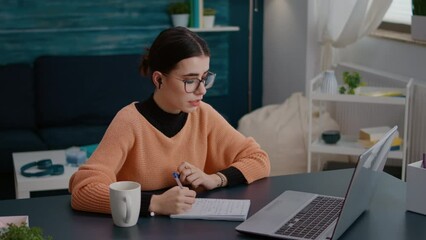 Female student attending online lesson on videoconference meeting, taking notes on textbook to study university knowledge. Woman using remote teleconference call for learning on laptop. - Powered by Adobe