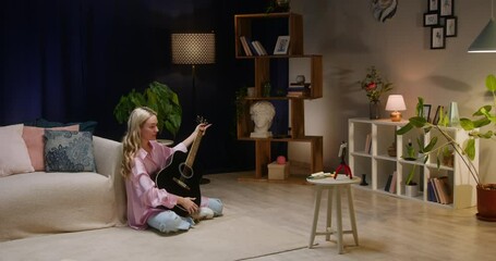 Woman playing acoustic guitar and recording video on smartphone camera in living room, video lesson. Female blogger making content for channel. Lifestyle vlog, talking at camera webcam online.