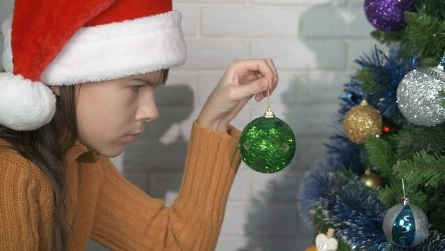 Sad teen in decorated Christmas living room. A teen girl with bad mood stay by the Christmas tree with green sparkling ball in hand.